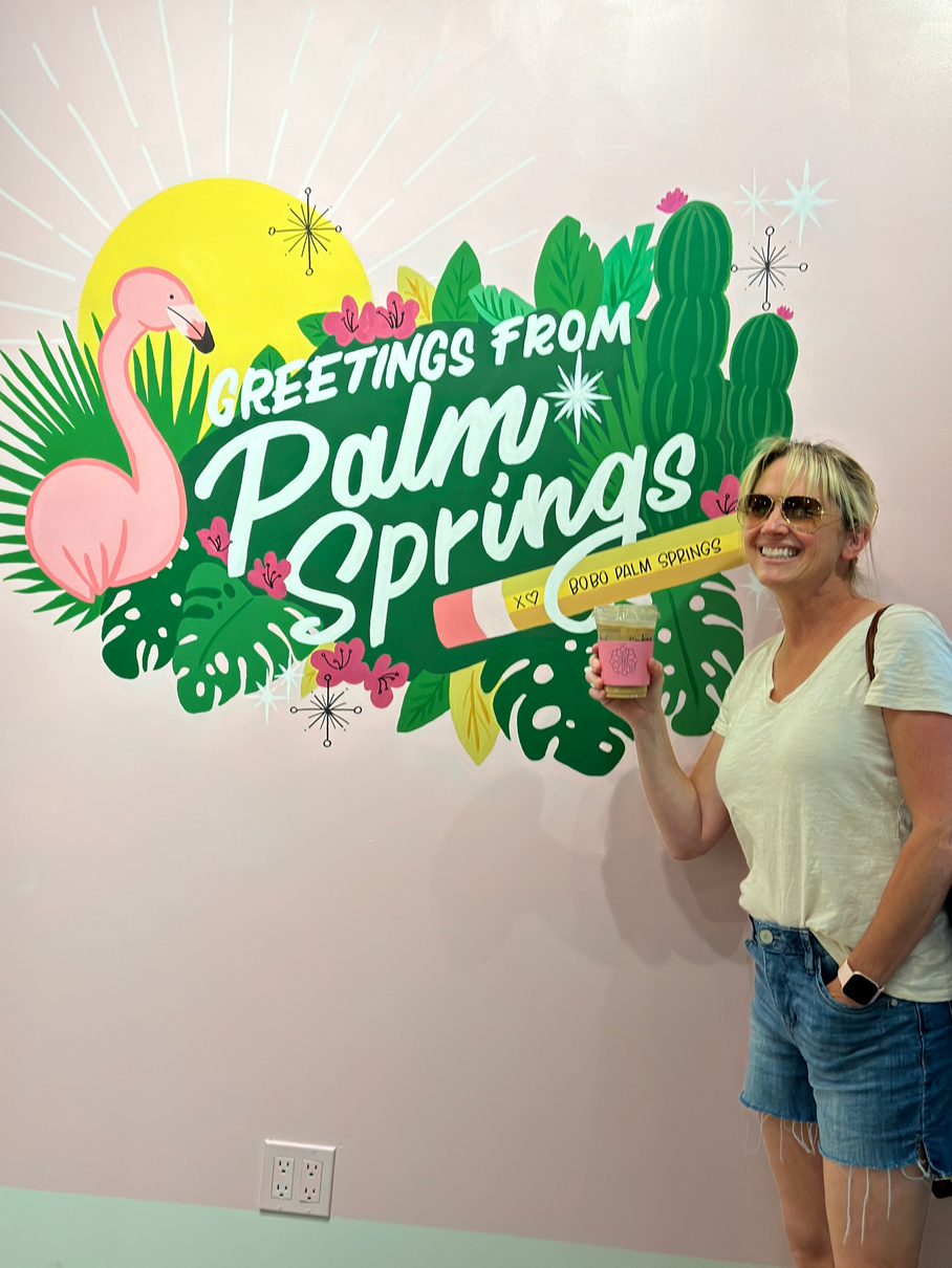 Lindsay Blake posing in front of the Greetings from Palm Springs mural at Bobo Palm Springs.